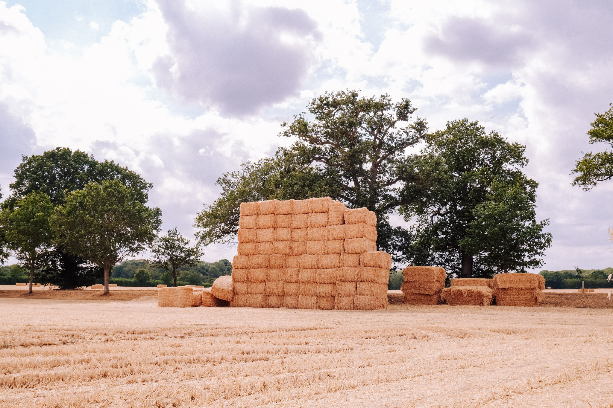 Stacked hay bales in a sunny field surrounded by trees under a partly cloudy sky.