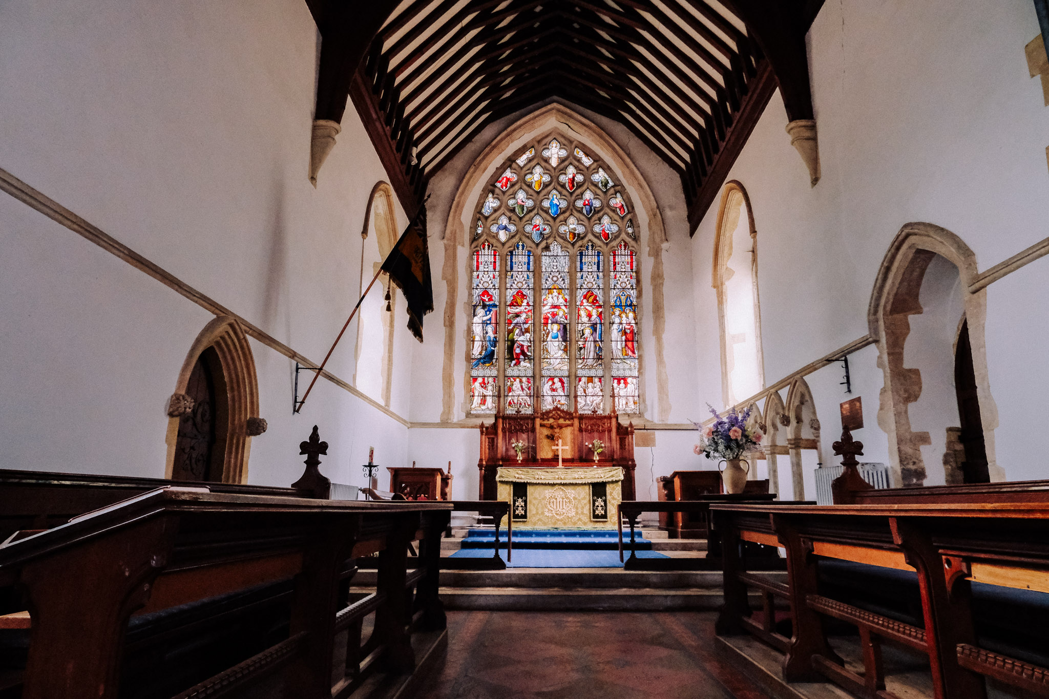 Interior view of a church showcasing wooden pews, an altar with a decorative cloth, and a large stained glass window in the background.