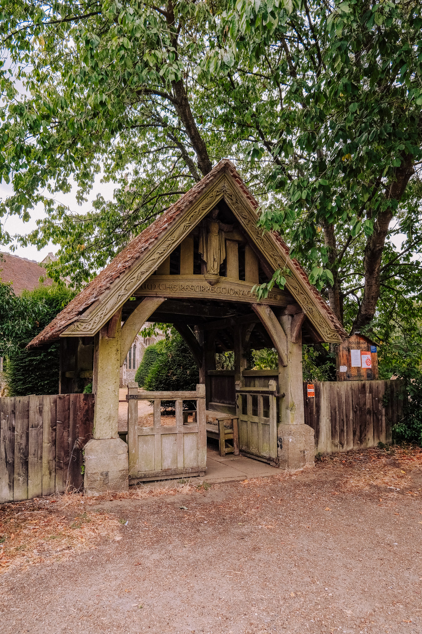 A rustic wooden structure with a triangular roof, featuring a statue and carved details, surrounded by trees and greenery.