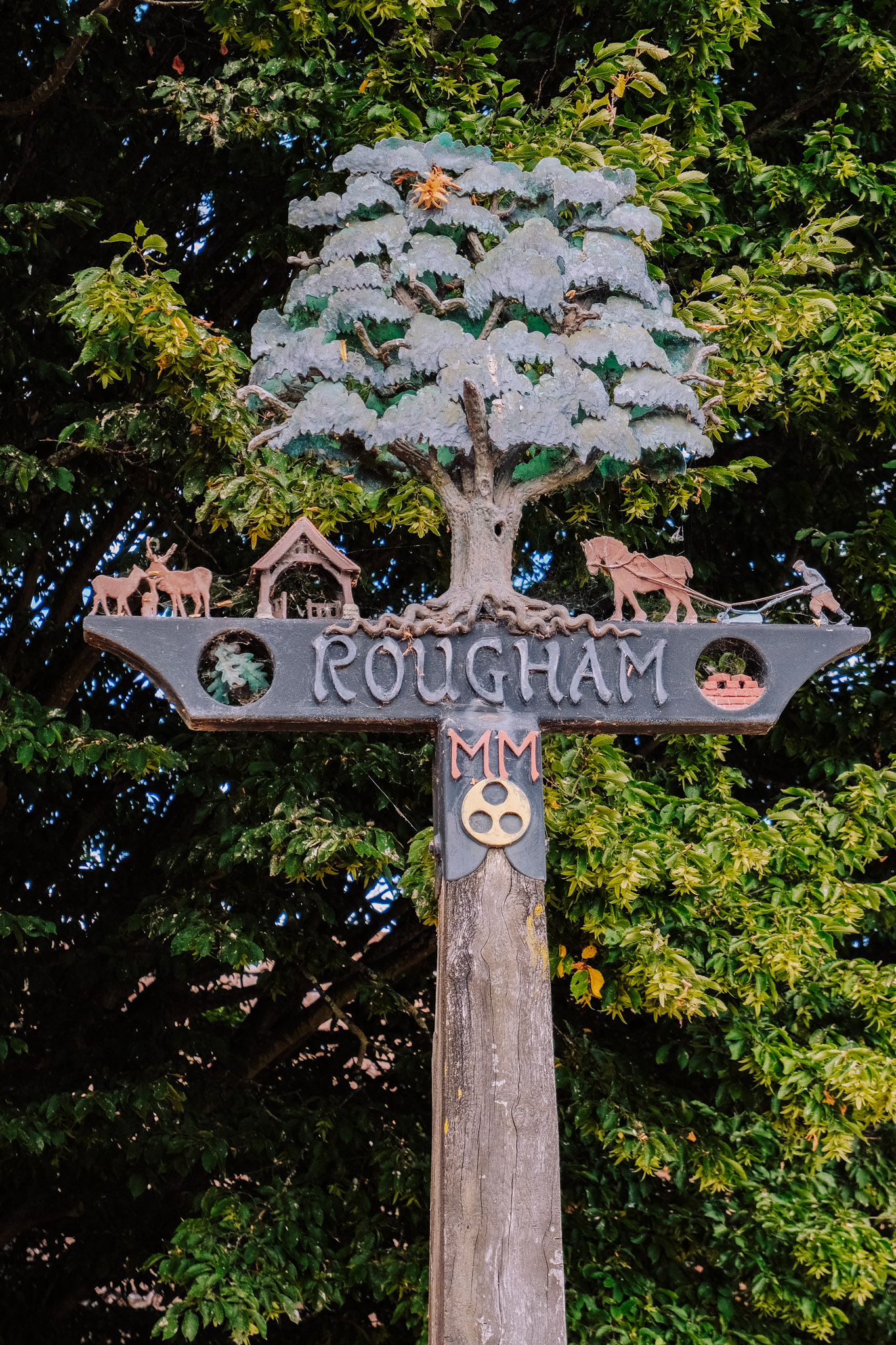 A decorative wooden sign for Rougham featuring a tree and various figures including animals and a house, surrounded by greenery.