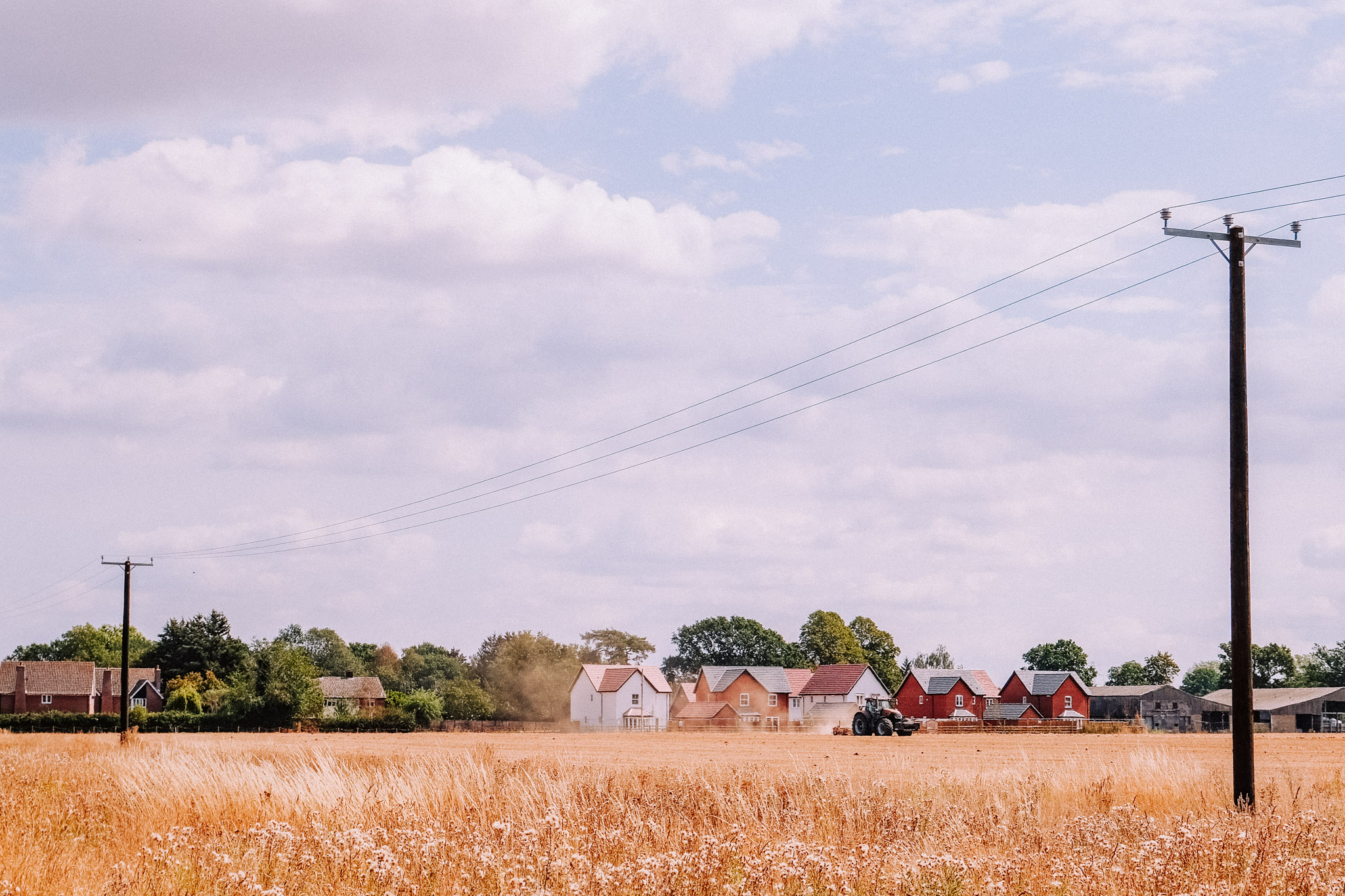 A wide view of a rural landscape featuring a golden field, power lines, and a tractor working in the distance, with houses partially visible among trees.