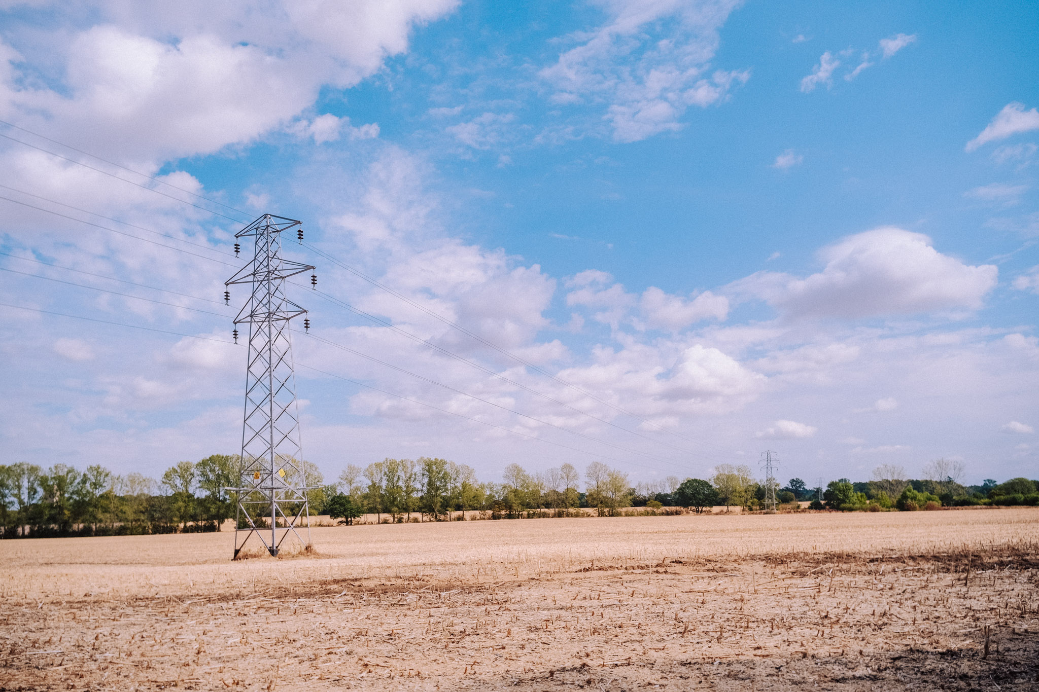 A tall electricity transmission tower surrounded by a dry field under a blue sky with scattered clouds and a line of trees in the background.