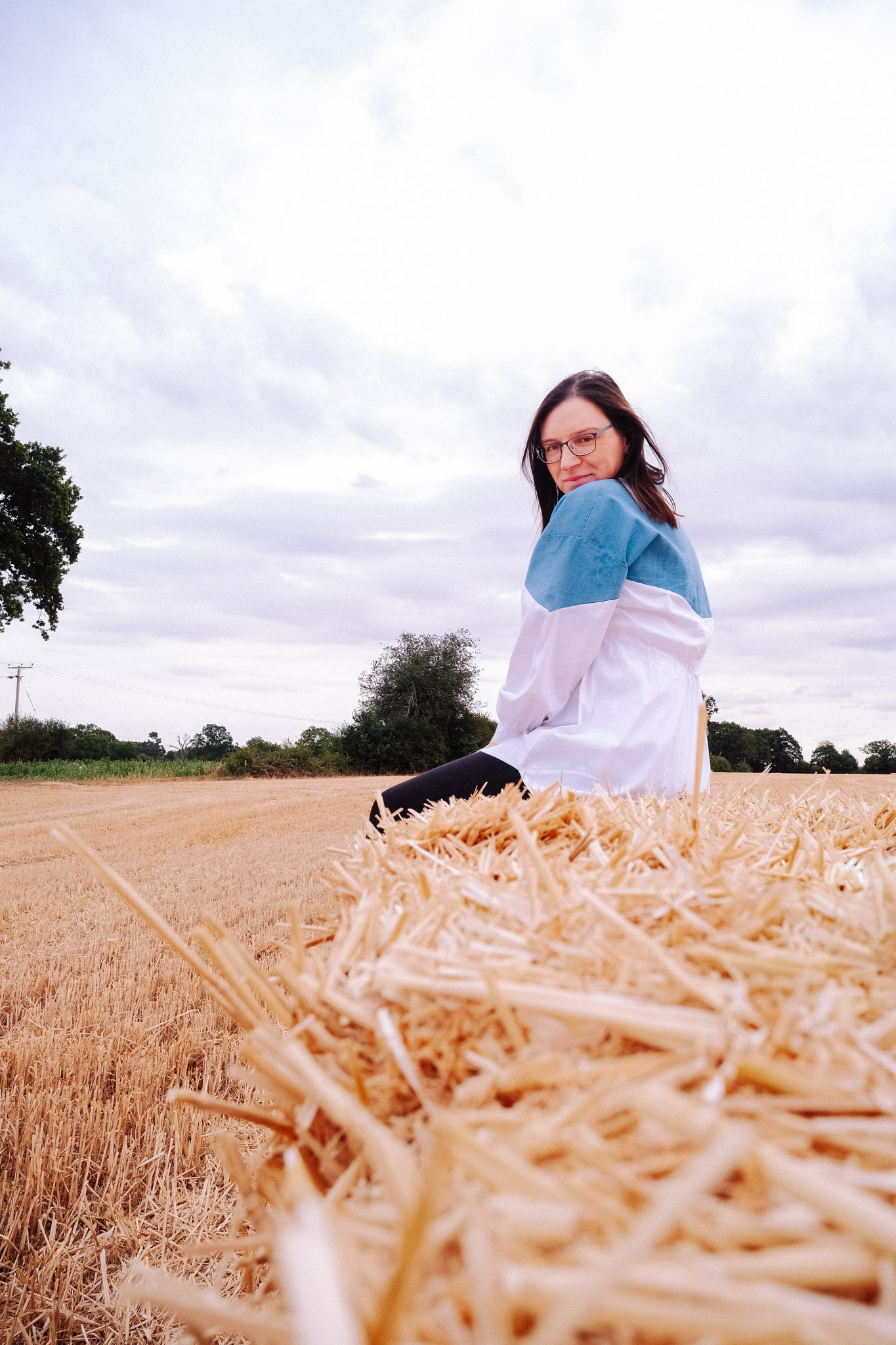 A woman sitting on a straw bale in a field, wearing a blue and white jacket, looking over her shoulder with a smile. The sky is partly cloudy and the landscape features green trees in the background.