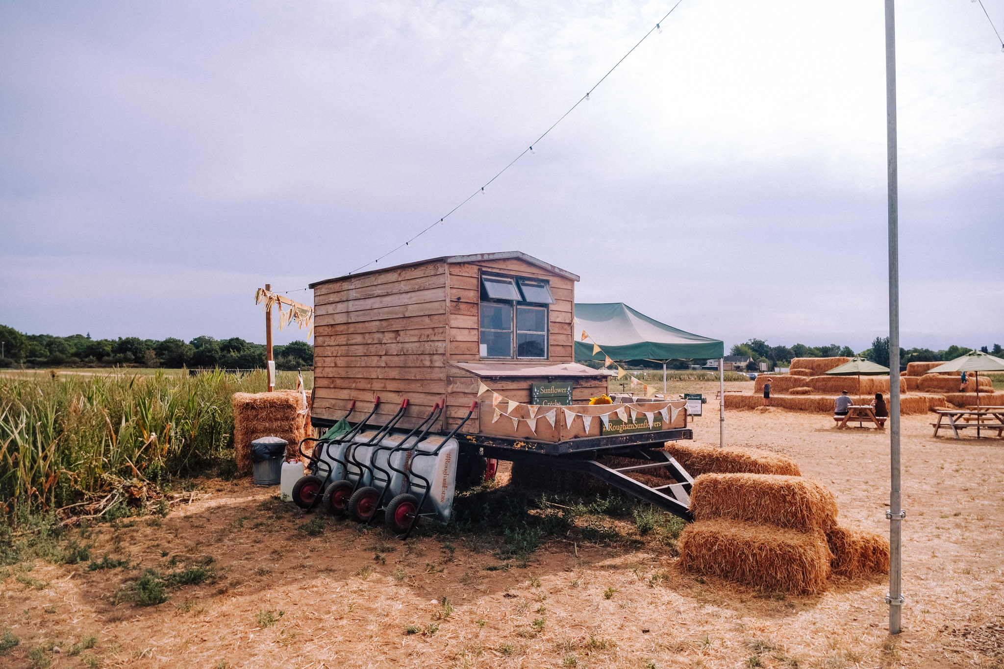 A wooden trailer displayed on a farm, featuring bunting decorations and a stall for selling sunflowers, surrounded by hay bales and open fields under a cloudy sky.