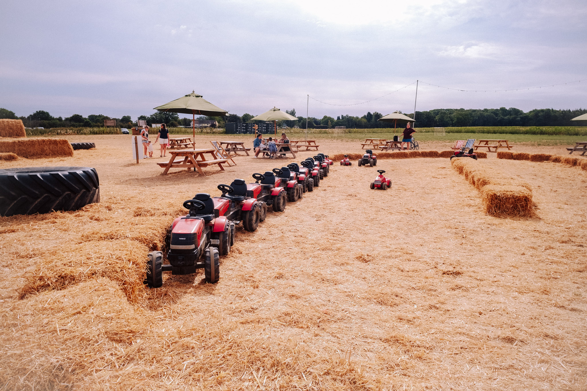 A child-friendly outdoor play area with toy tractors arranged in a line on a straw-covered ground, surrounded by hay bales and picnic tables under umbrellas.