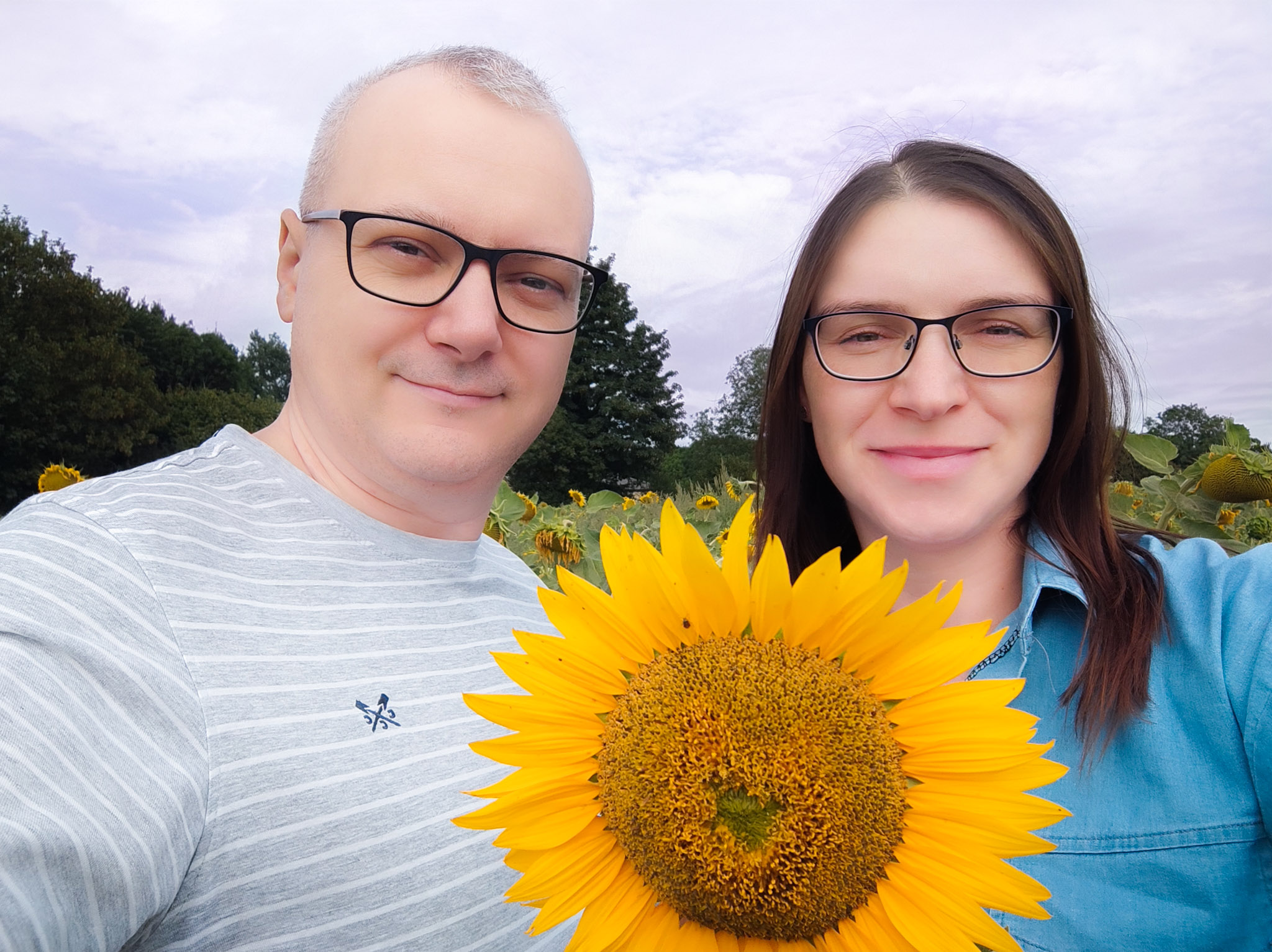 A man and a woman smiling together while holding a large sunflower, surrounded by a field of sunflowers under a cloudy sky.