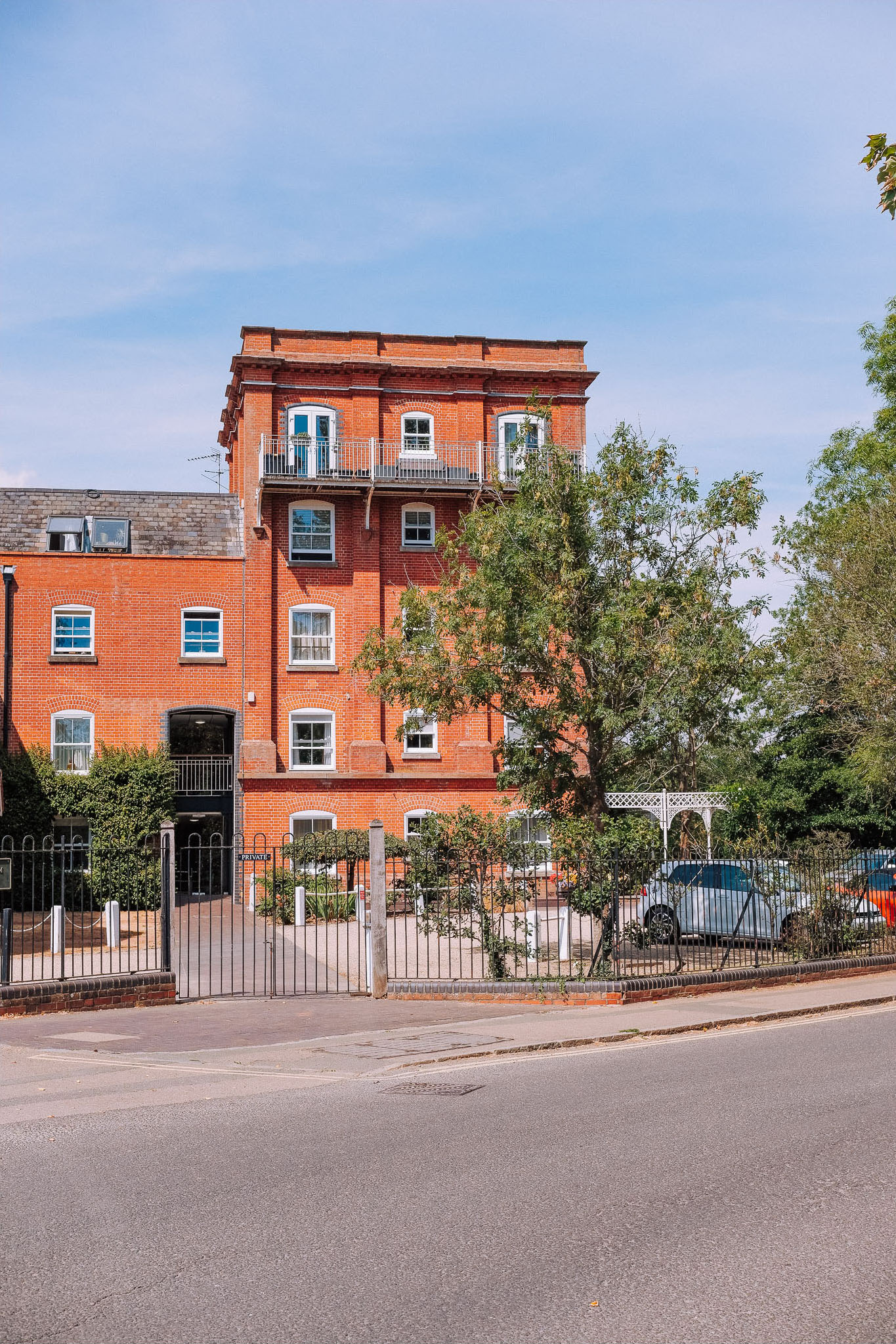 Red brick building with multiple windows and a balcony, surrounded by trees and a gated entrance, located at the corner of a street.