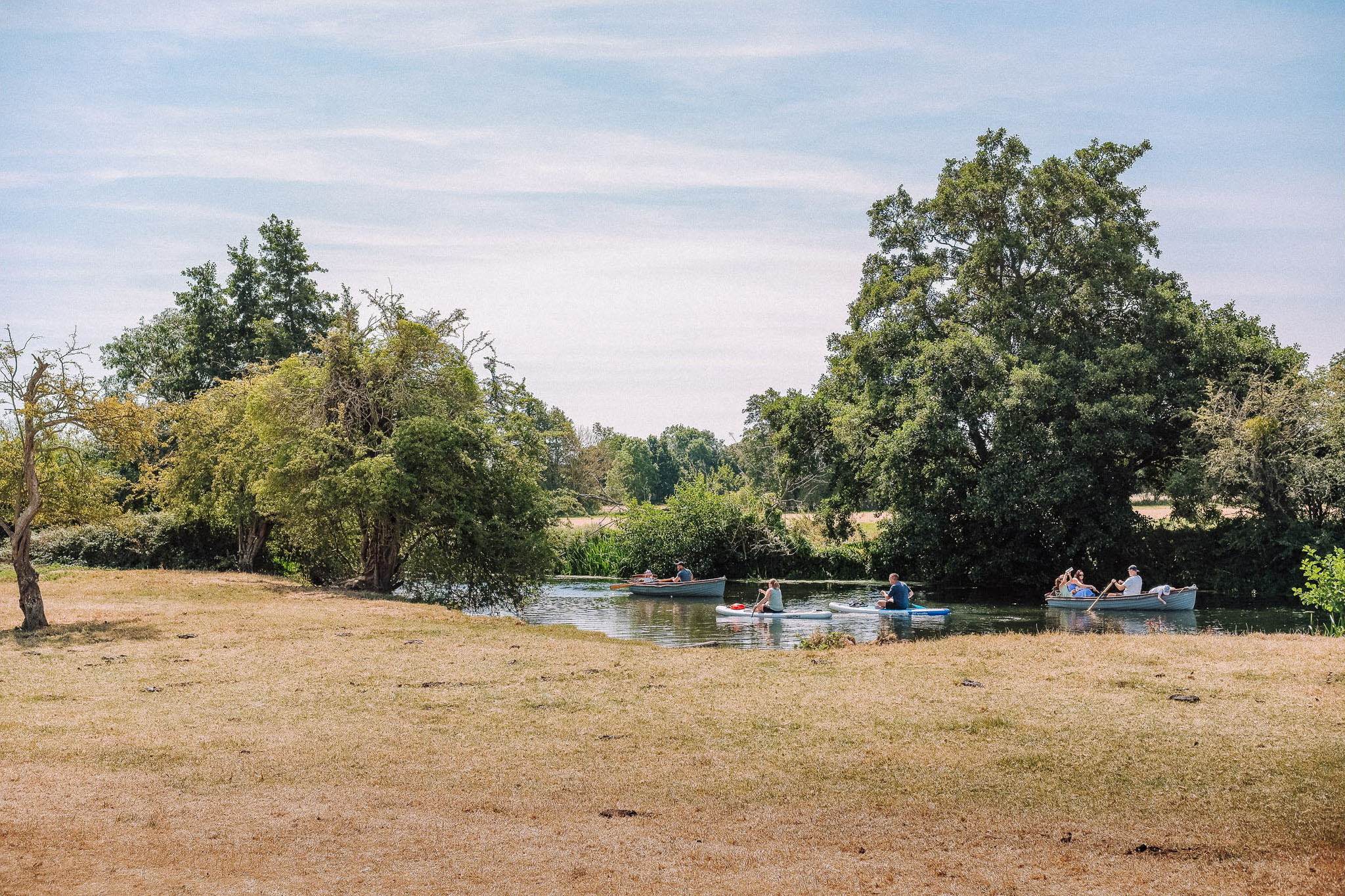 A scenic view of a calm river surrounded by lush greenery, with several people in small boats enjoying a leisurely paddle on a sunny day.