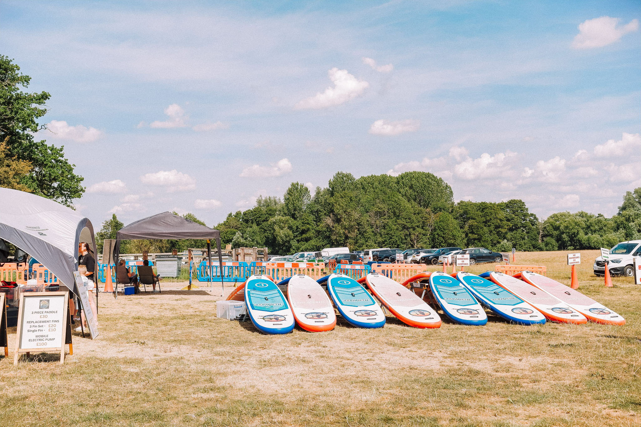 A sunny outdoor scene featuring several stand-up paddleboards neatly arranged on grass, with a couple of tents and a parking area in the background.
