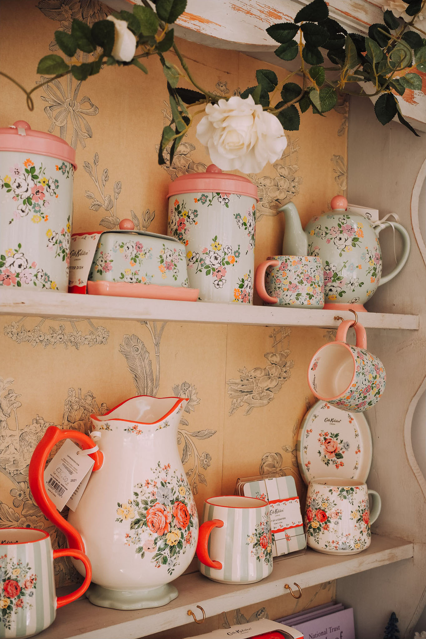 A collection of floral-patterned kitchenware arranged on a shelf, including teapots, mugs, and storage containers, against a vintage wallpaper backdrop.