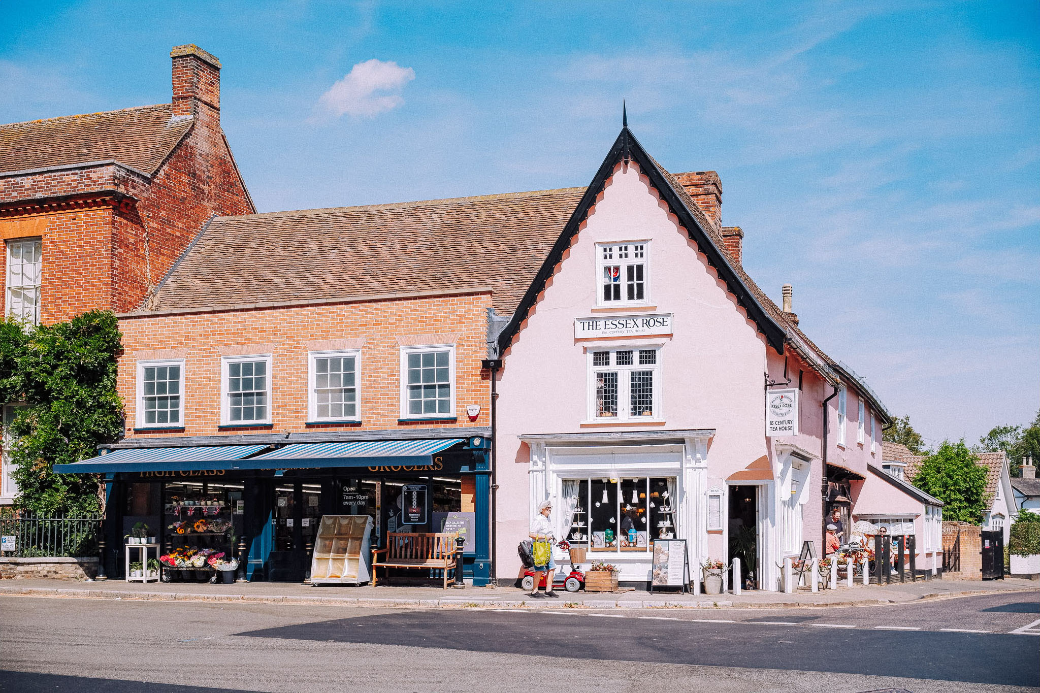 A quaint street scene featuring two shops: a grocery store with a blue awning and a pink building labeled 'The Essex Rose', set against a blue sky.