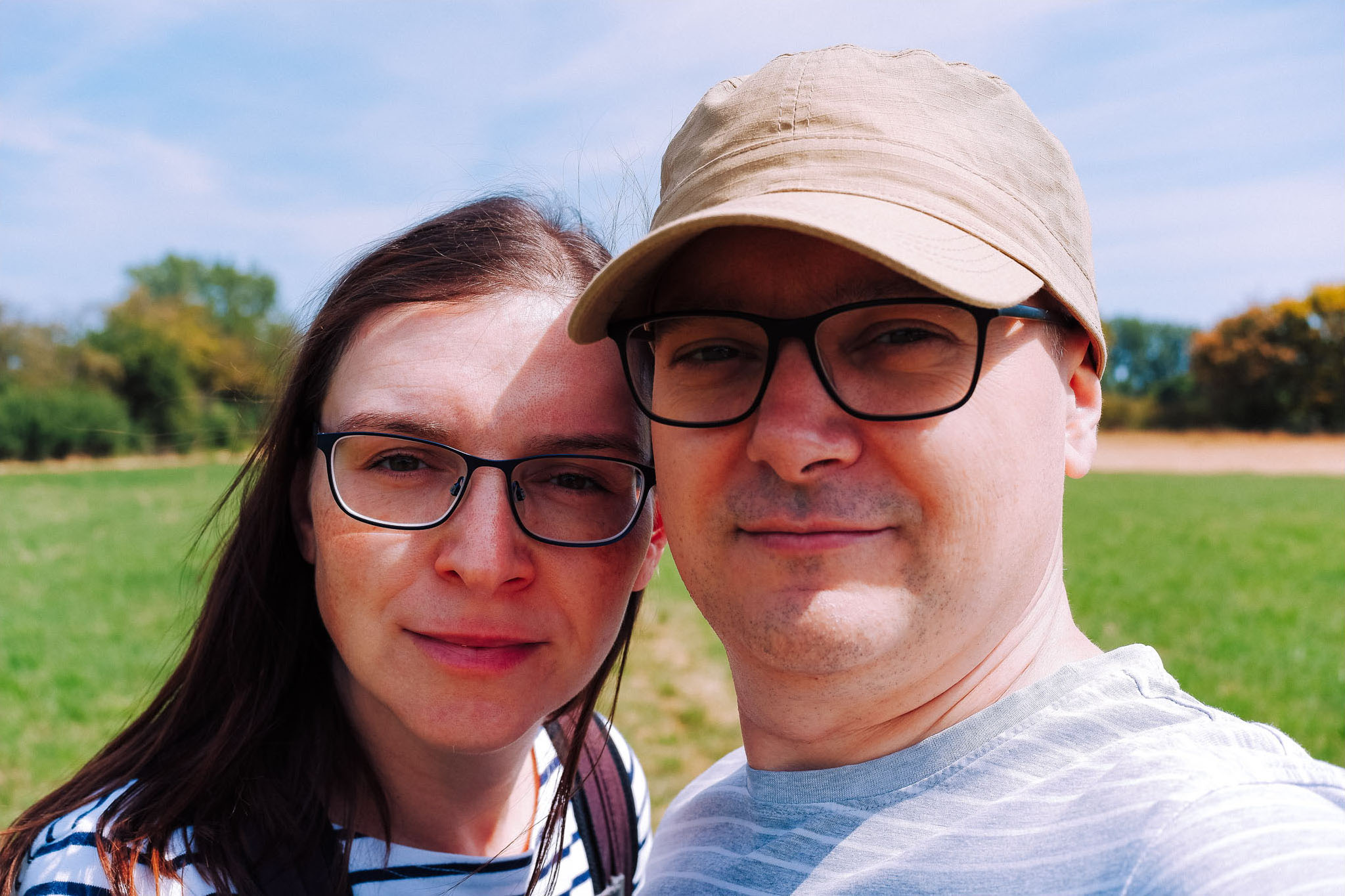 A couple taking a selfie outdoors in a green field, featuring a woman with long brown hair and glasses, and a man wearing a cap and glasses, with a clear blue sky in the background.