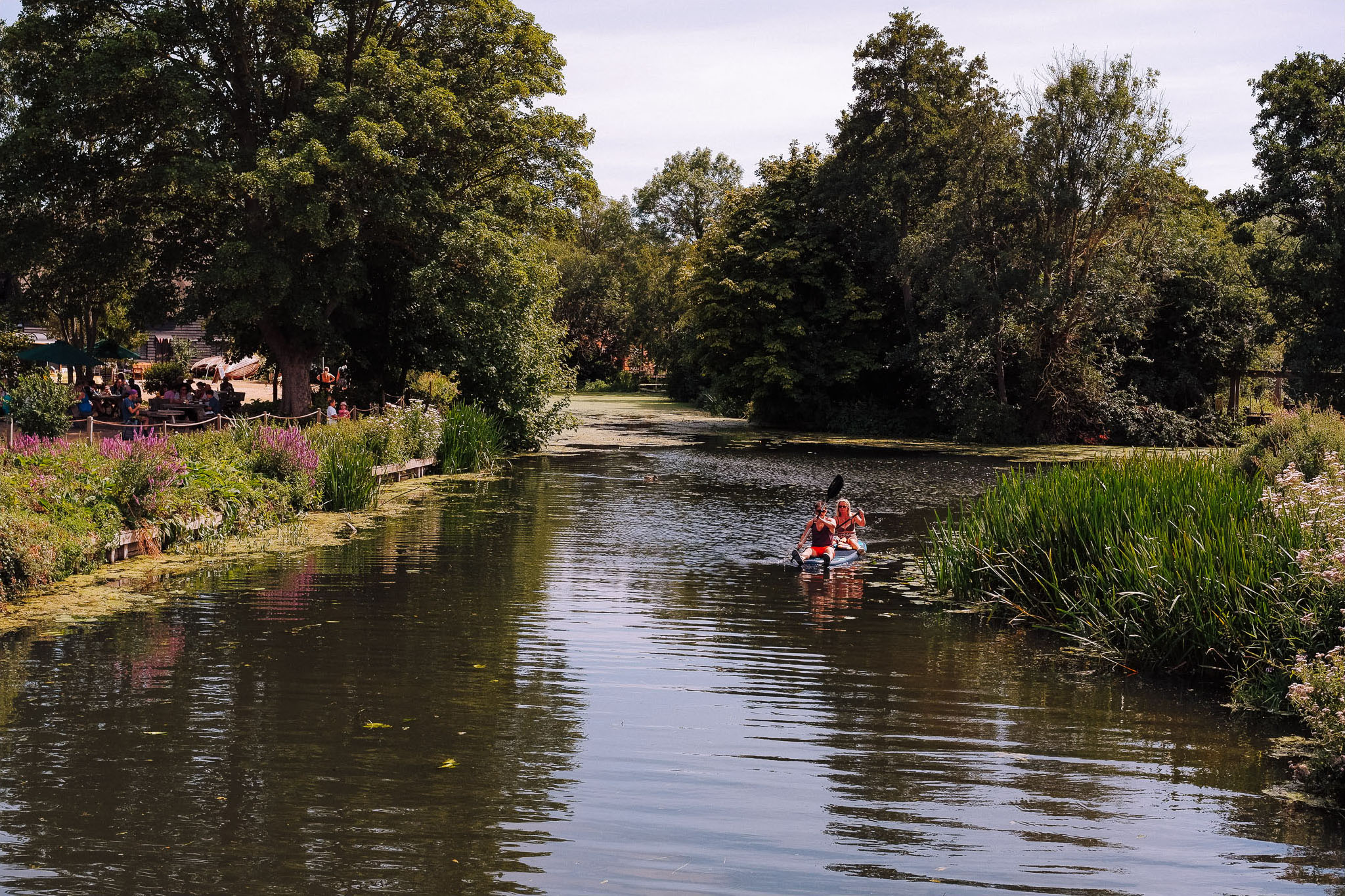 Two people kayaking on a tranquil river surrounded by lush greenery and flowering plants, with a visible seating area in the background.