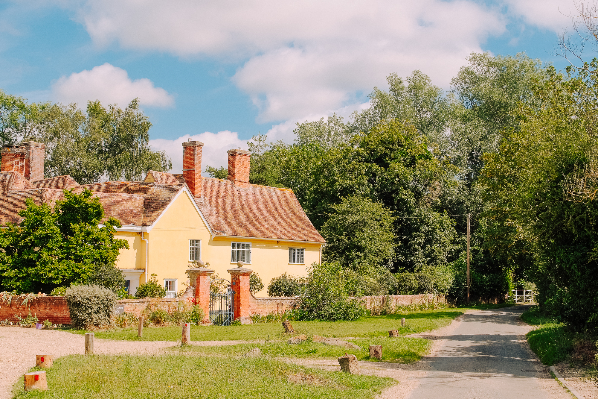 A picturesque yellow house surrounded by lush greenery, with a gravel path leading to the road. The sky is bright with scattered clouds.