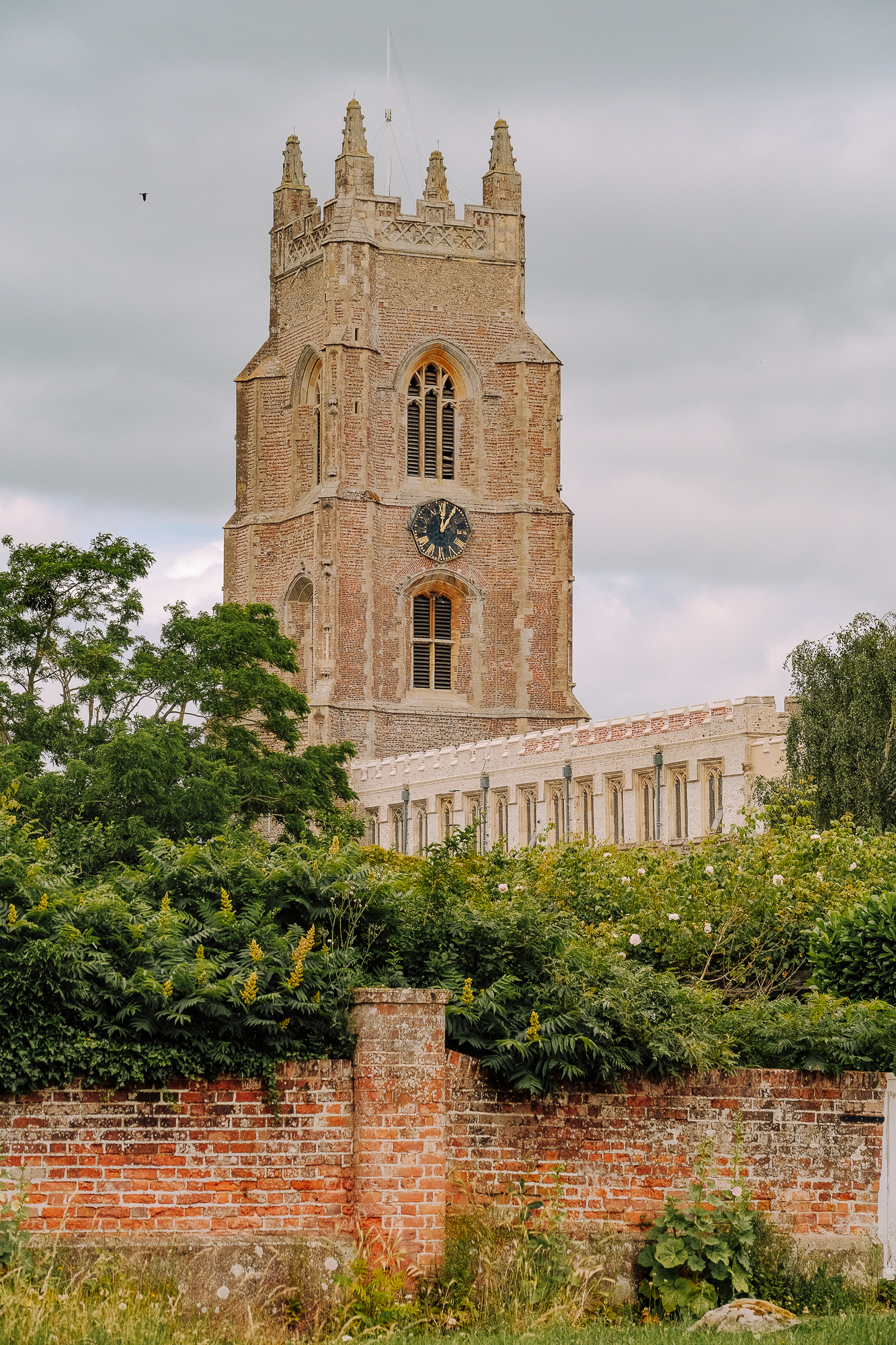 A historic brick church tower with a clock, surrounded by lush greenery and a brick wall in the foreground under a cloudy sky.