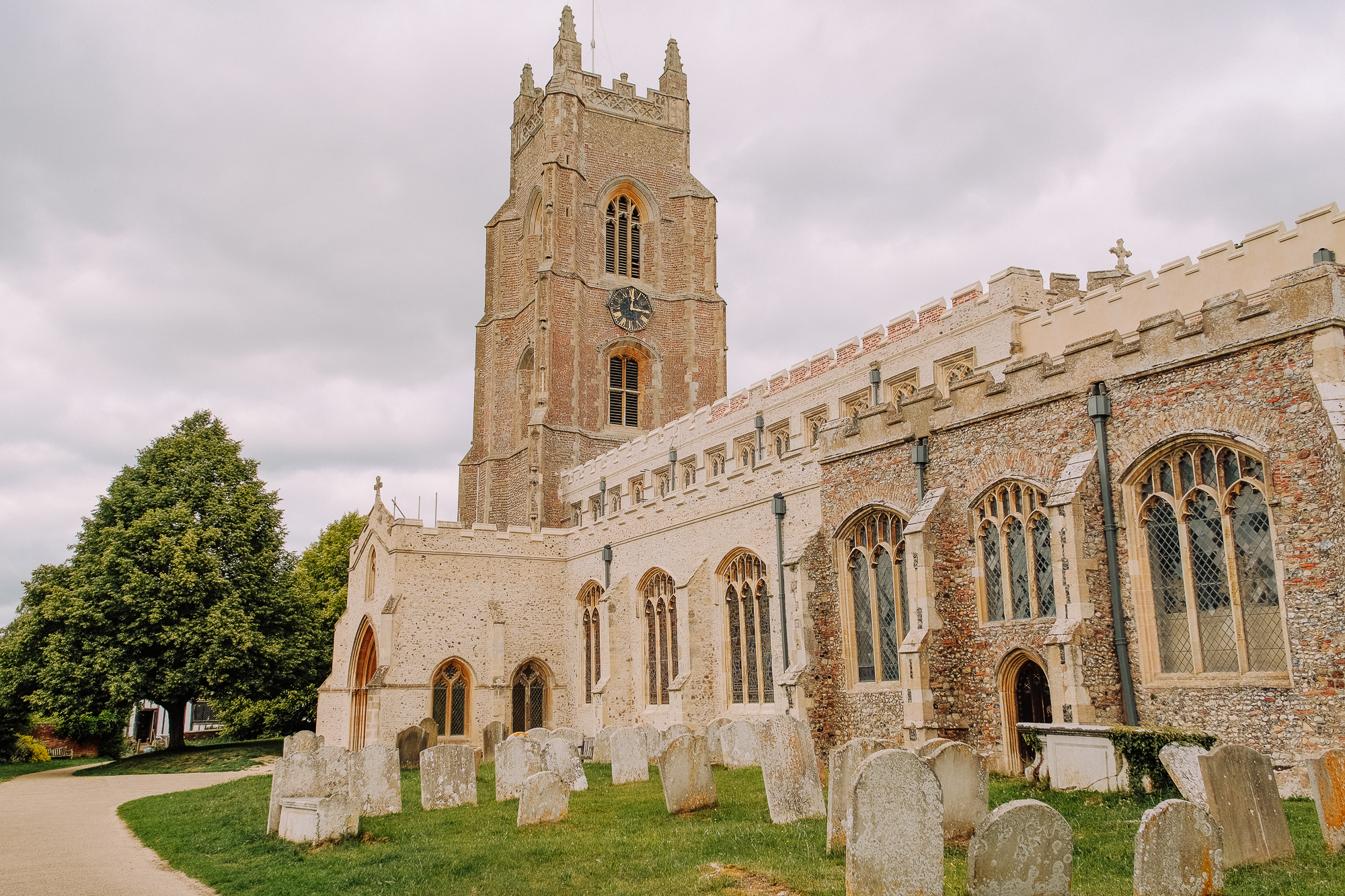 Historic church exterior featuring a tall bell tower and intricate stonework, surrounded by gravestones and lush greenery under a cloudy sky.
