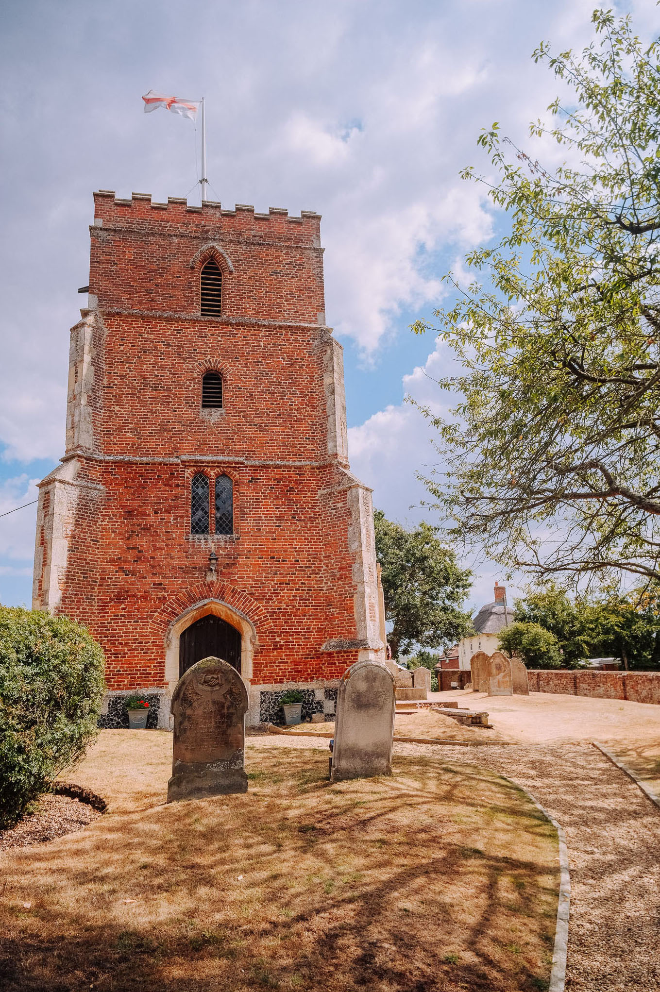 A historic brick church tower with a flag, surrounded by gravestones and a pathway in a churchyard, under a partly cloudy sky.