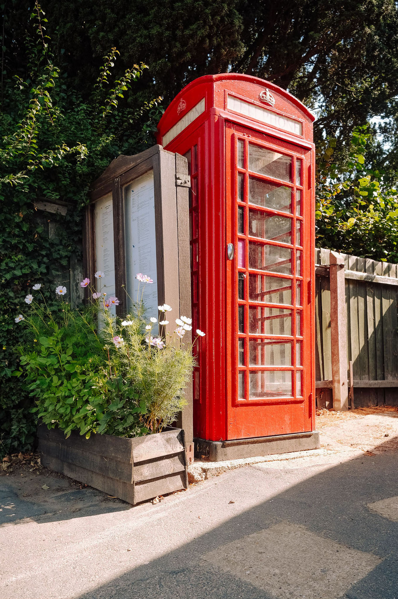 A traditional red British telephone box next to a wooden notice board and surrounded by lush greenery and blooming flowers.