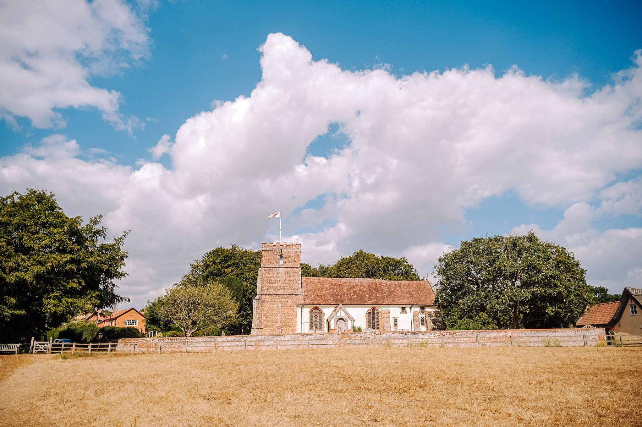 A countryside scene featuring a historic church with a tall tower and a flag, surrounded by trees and a dry field under a cloudy blue sky.