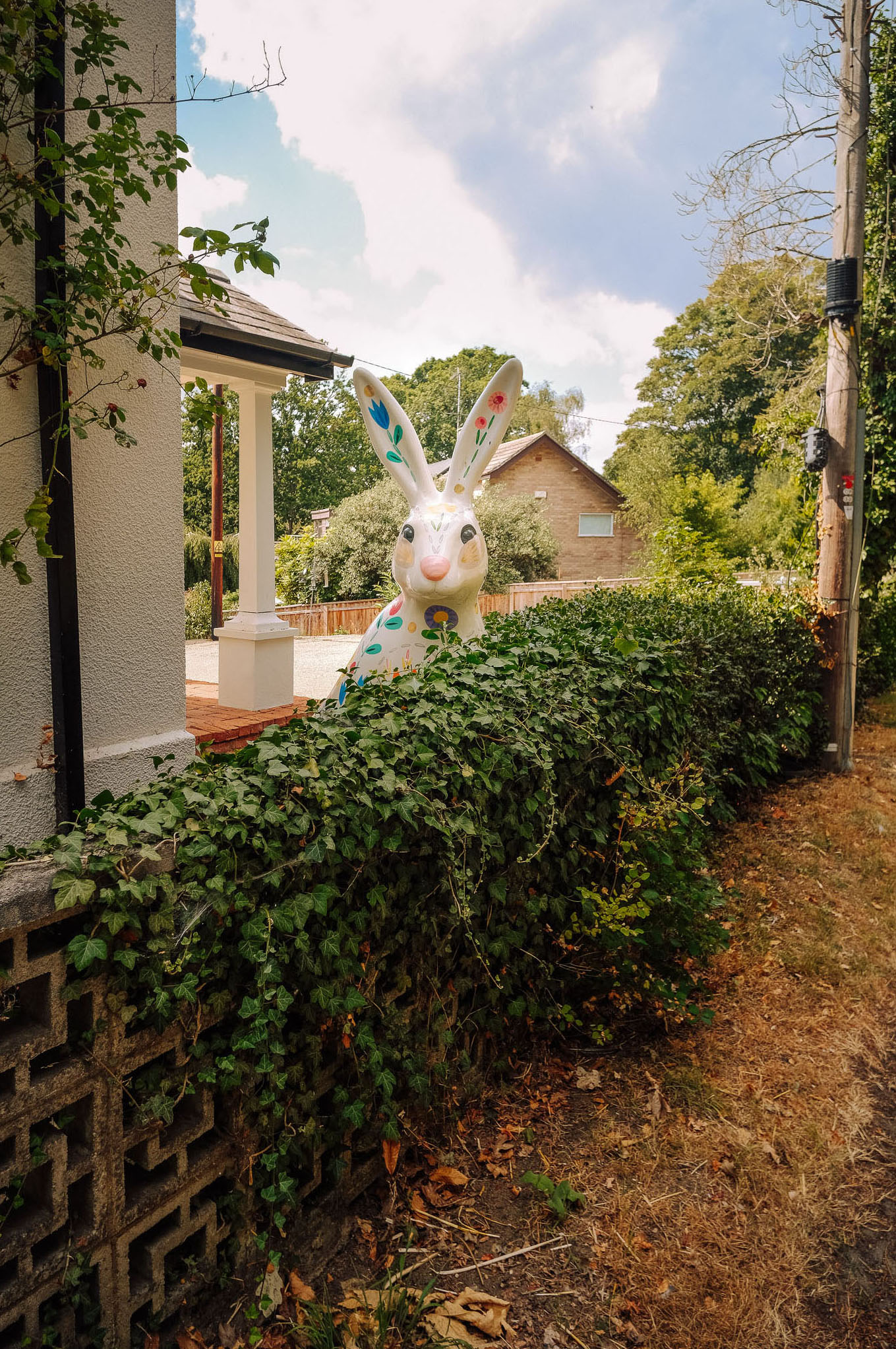 A large, colorful bunny figure peeking over a hedge near a residential area, with a house and trees in the background.