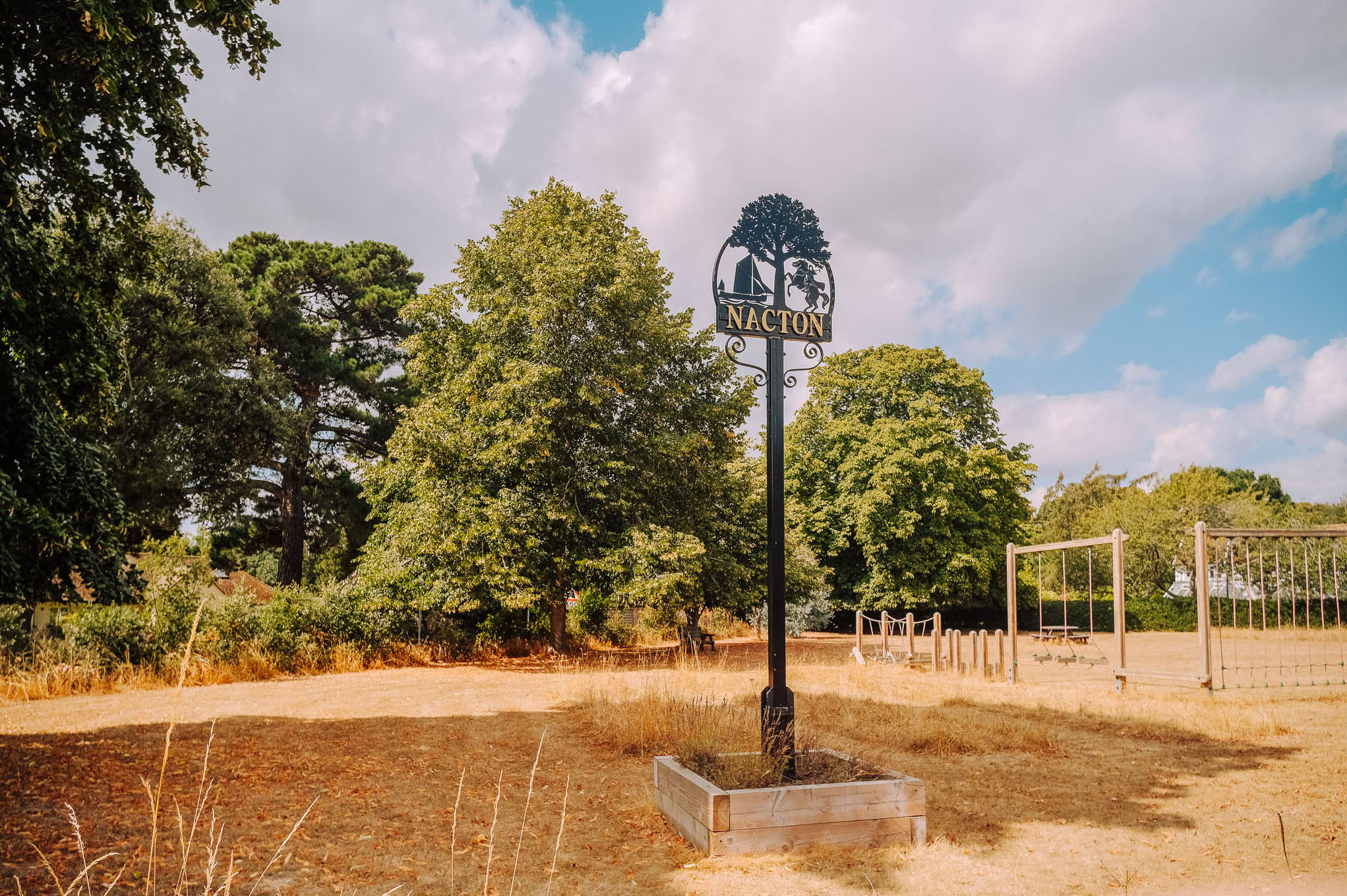 A sign post for Nacton surrounded by trees and a grassy area under a cloudy sky.