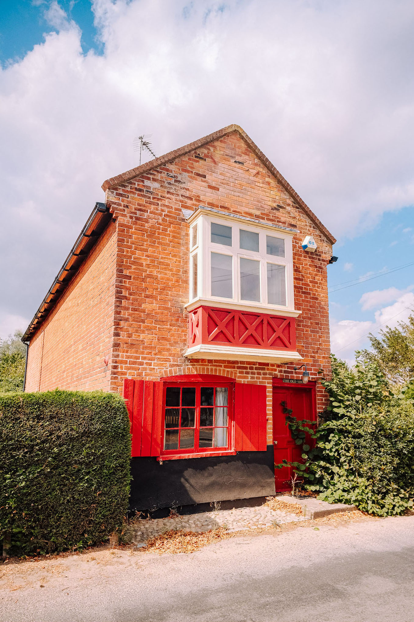 Charming brick house with red accents, featuring a front balcony and a lush green hedge.