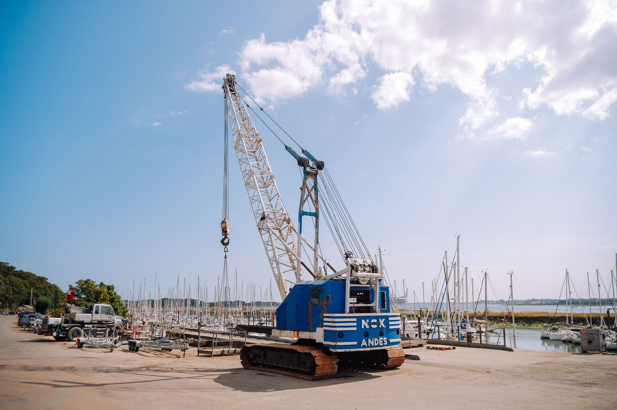 A blue and white crawler crane named Andes parked at a marina, with several sailboats and a clear blue sky in the background.