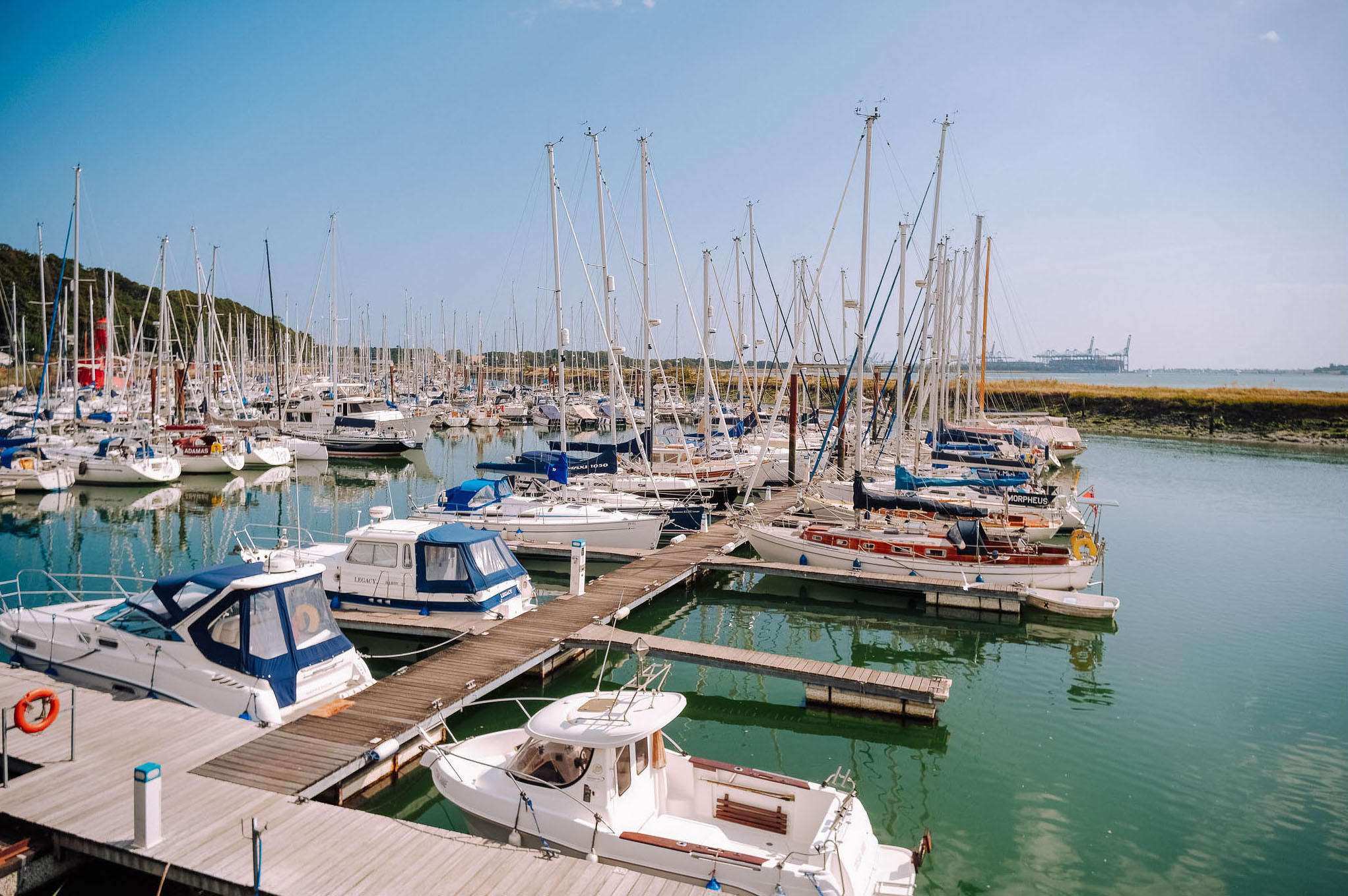 A busy marina filled with various boats and yachts, with masts silhouetted against a clear blue sky. The water reflects the boats, and wooden docks extend through the marina.