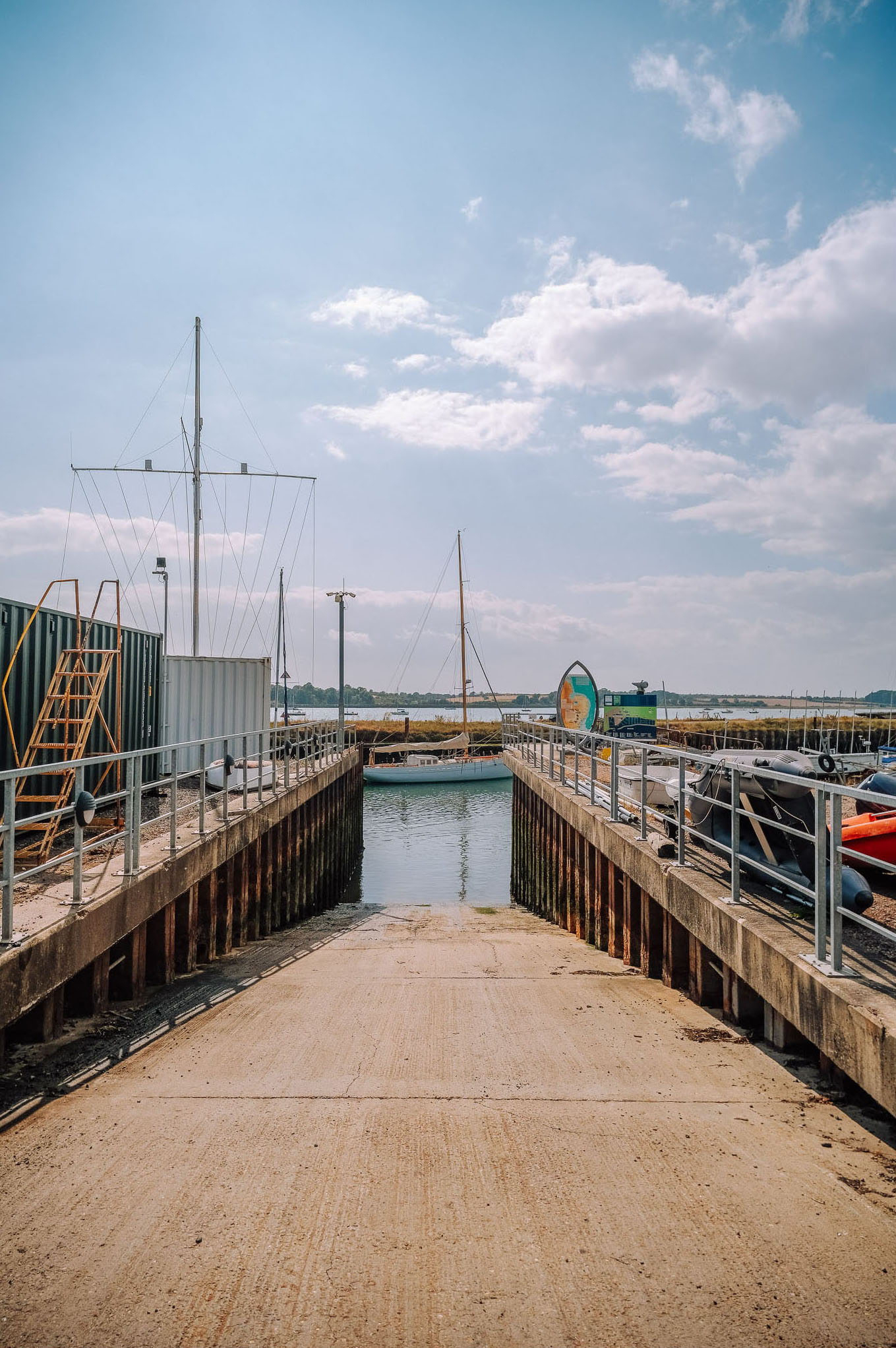 A view of a marina with a concrete ramp leading down to the water, surrounded by moored boats and a clear blue sky with scattered clouds.