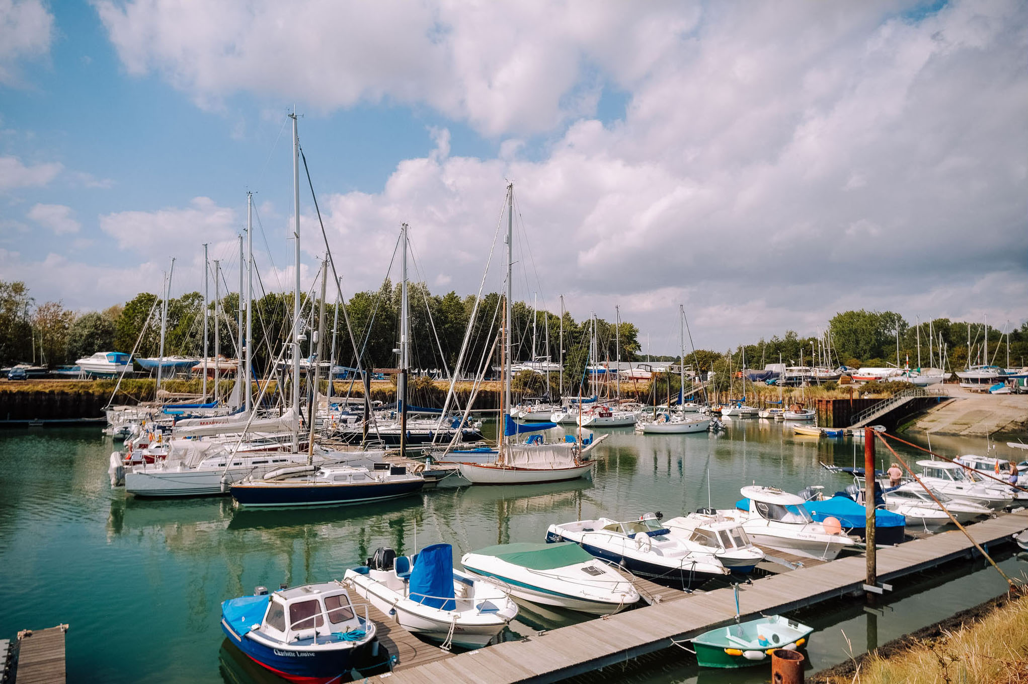 A view of a marina filled with various sailboats and motorboats, surrounded by green trees and a partly cloudy sky.