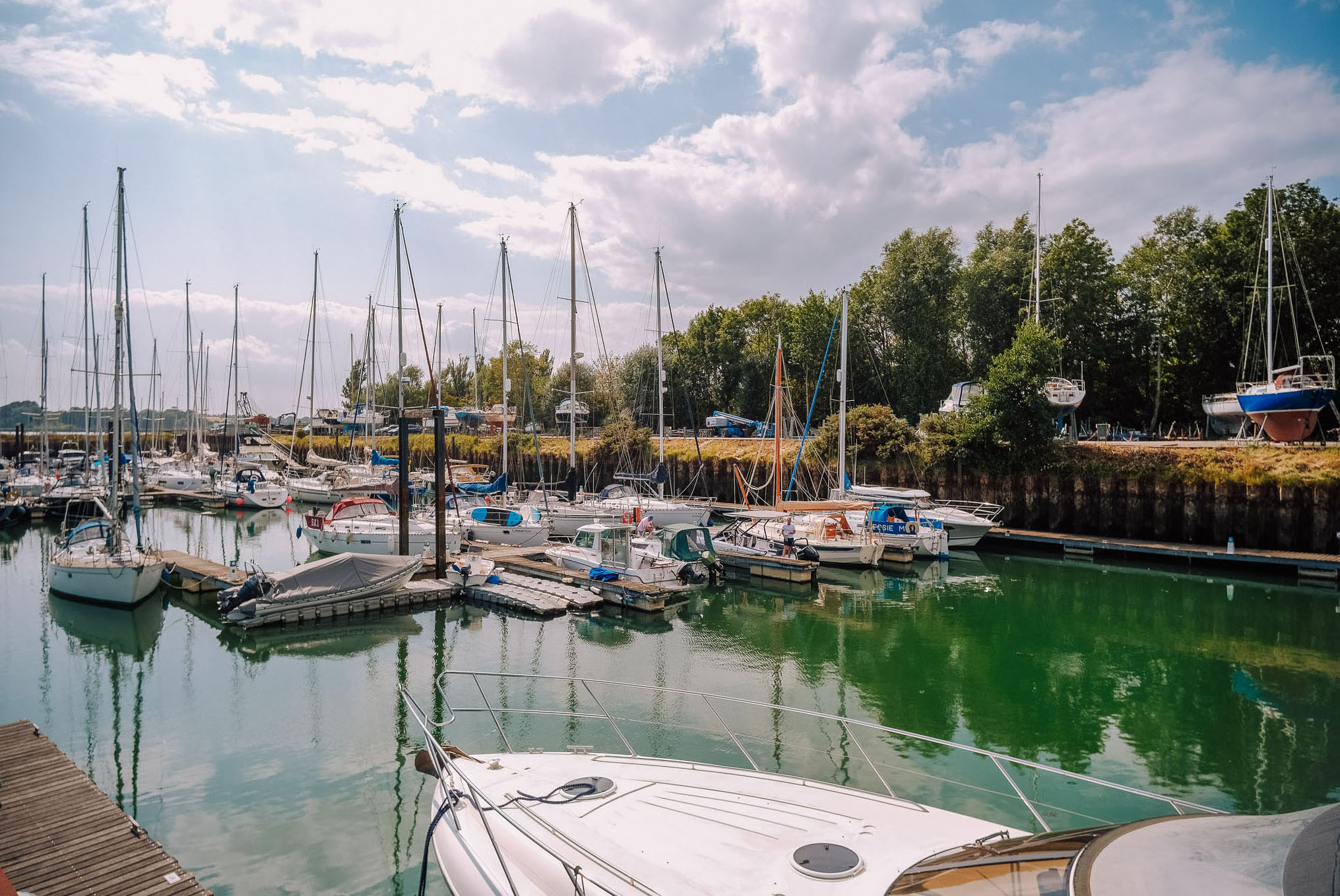A scenic marina with numerous sailboats docked in calm green waters, with cloudy skies and foliage in the background.