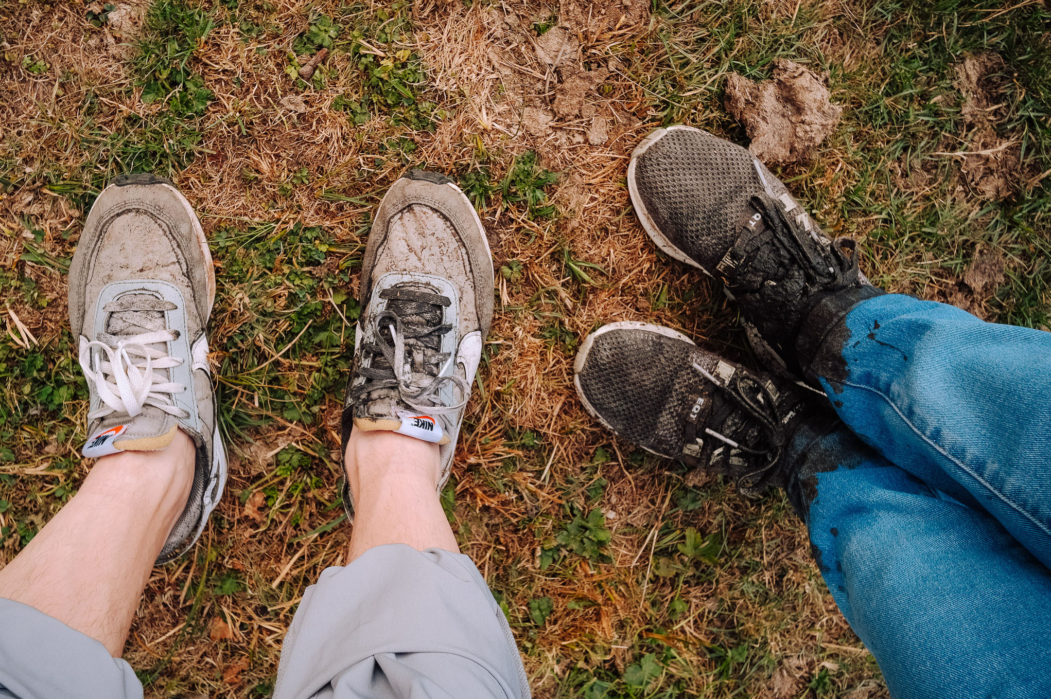 Close-up of two pairs of dirty sneakers on grass, one pair grey and white, the other black, with muddy jeans visible.